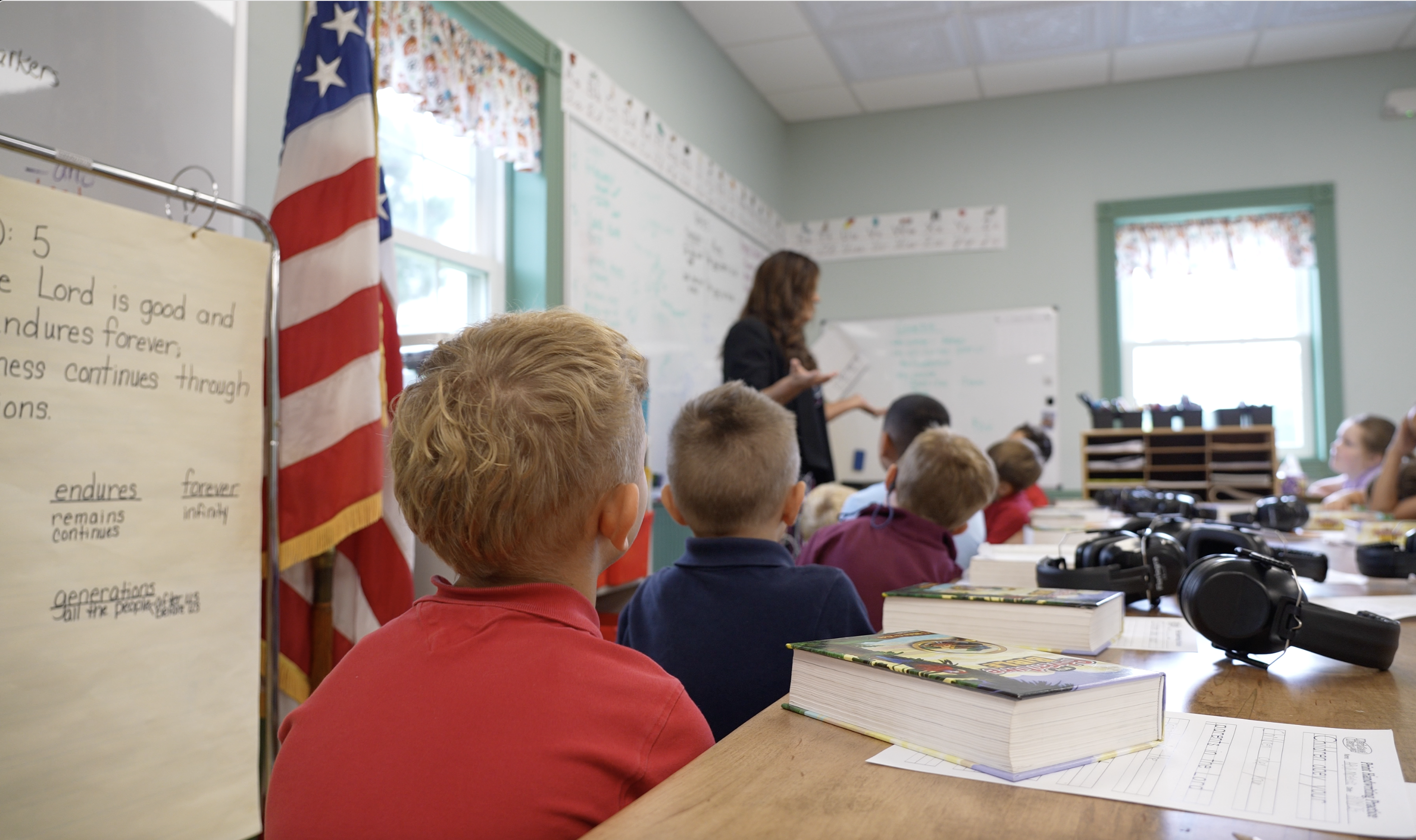 Dennita teaching a classroom of children at The Little Red Schoolhouse — the Bible verse on the flip chart reads 'The Lord is good and endures forever'